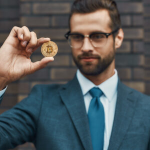 Digital currency. Handsome businessman in formal wear and eyeglasses holding bitcoin in one hand and looking at it while standing against brick wall