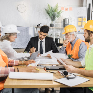 Multiracial men and women in formal outfit and helmets sitting at table with lots of blueprints